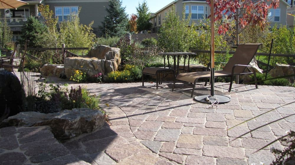 Landscaped backyard with stone patio, lounge chairs, rock feature, and colorful flowers.