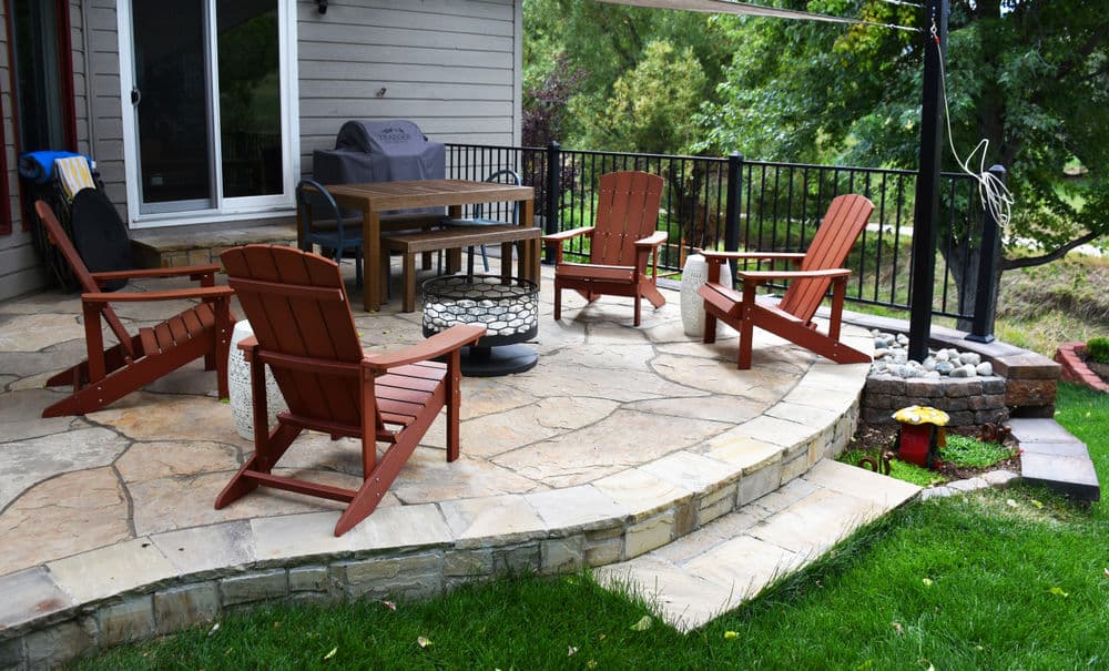 Cozy patio with four Adirondack chairs and a fire pit surrounded by greenery.