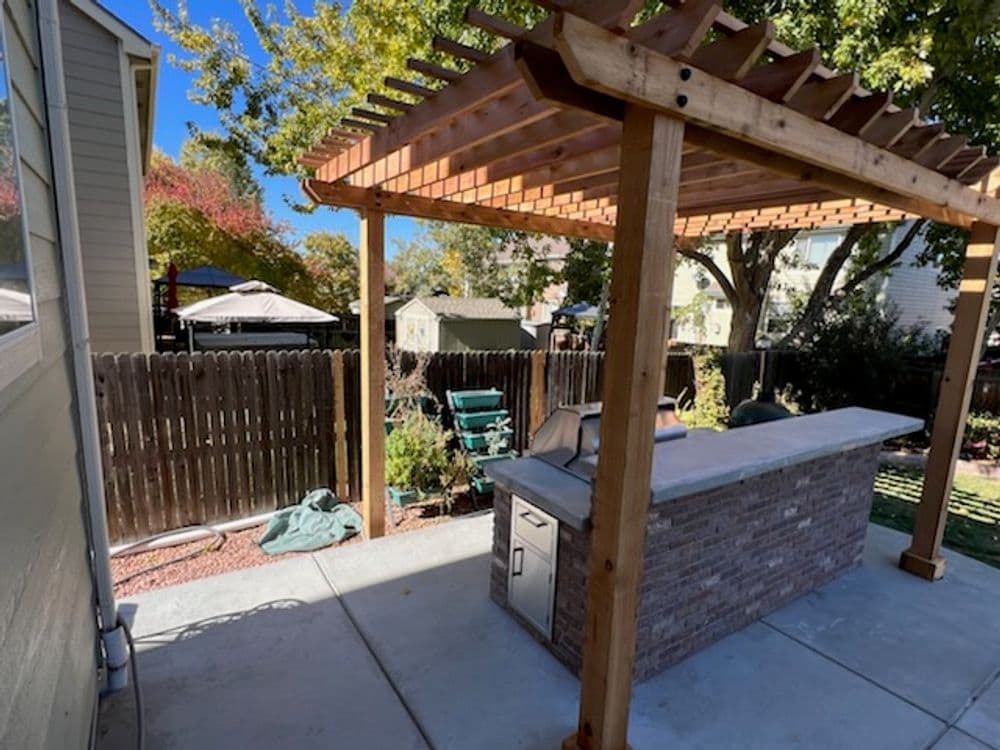 Backyard patio with wooden pergola, outdoor kitchen, and green lawn under clear blue sky.
