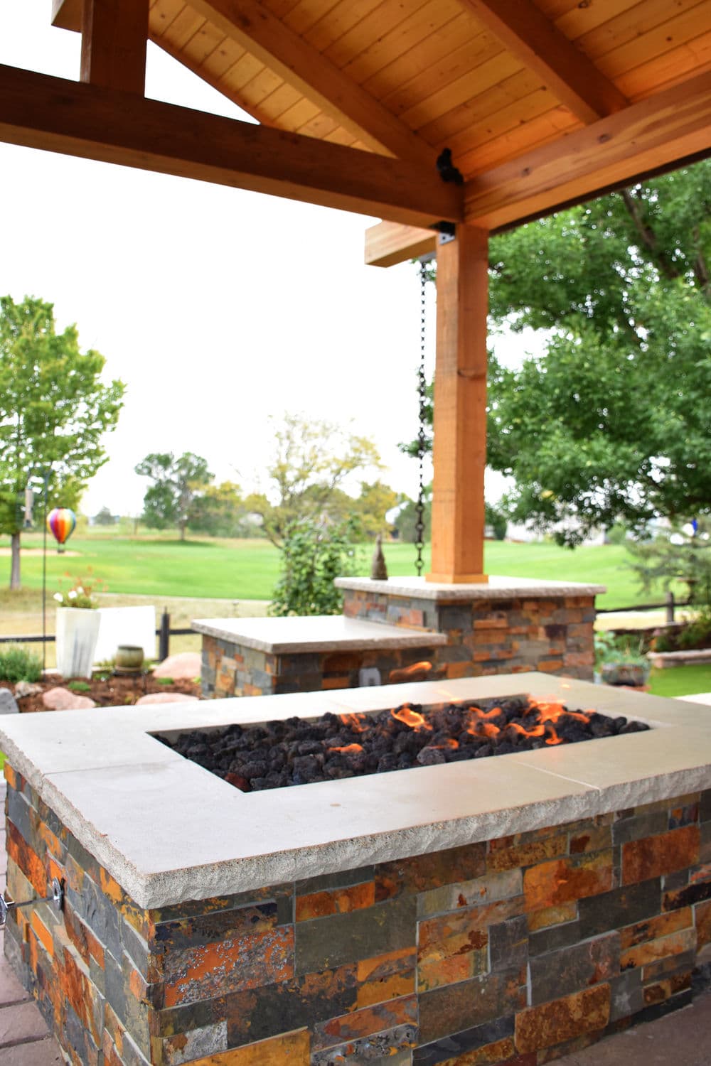 Modern outdoor fire pit with stone surround under a wooden pergola, overlooking a green landscape.