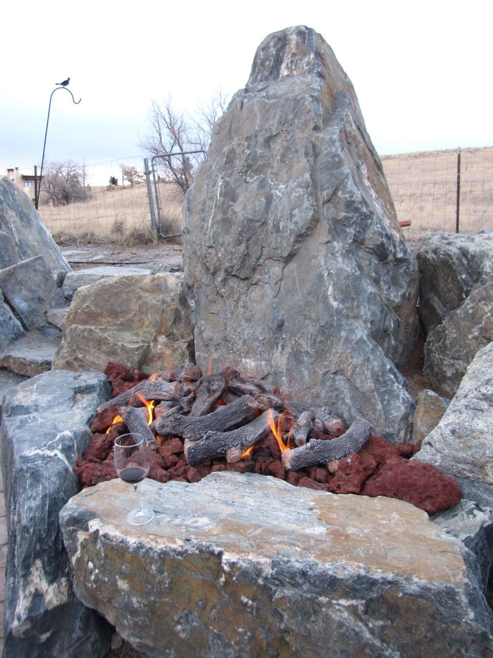 Outdoor fire pit with flames, surrounded by large rocks and a glass of red wine nearby.