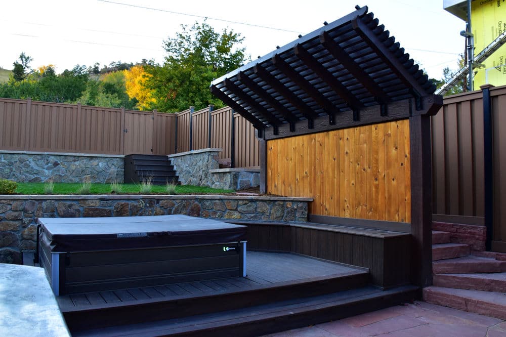 Modern wooden hot tub in a landscaped backyard with stone wall and pergola.