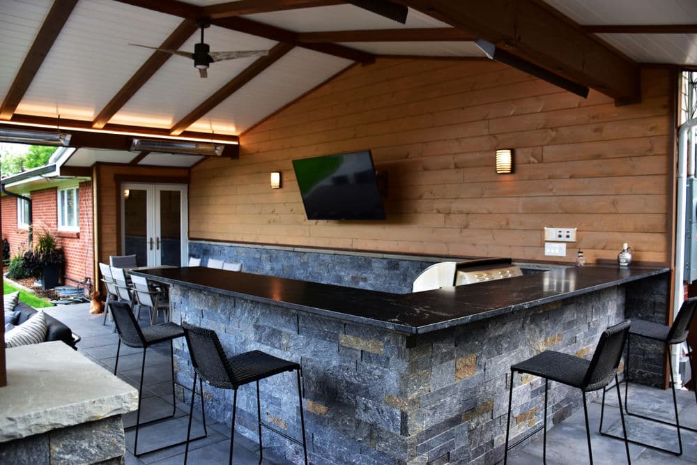 Outdoor bar area with stone countertop and modern seating, featuring a TV and vaulted ceiling.